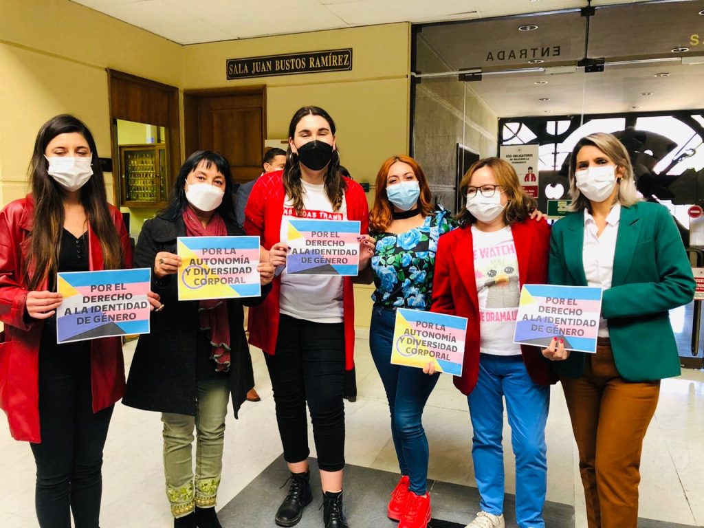 Emilia Schneider junto a activistas y parlamentarias sosteniendo carteles a favor del derecho a la identidad de género, usando mascarillas.