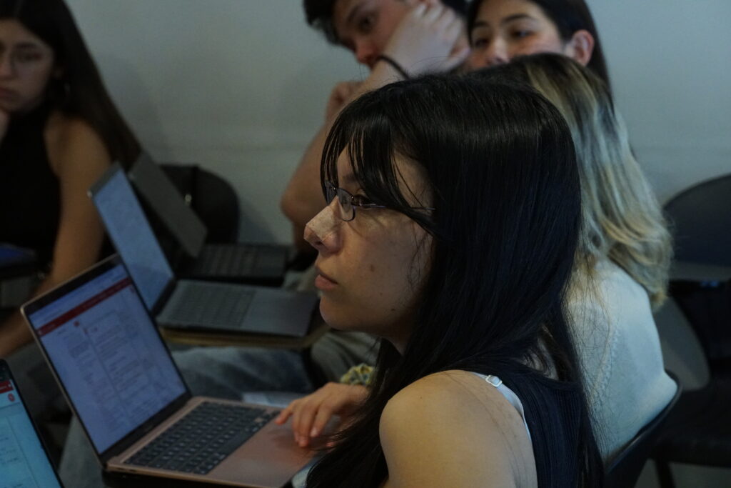 Estudiante en primer plano mientras escucha la clase de Lorena Lorca y detrás se ven a otros estudiantes con sus computadores,