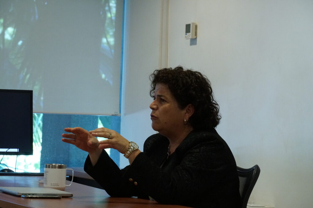Abogada Lorena Lorca de perfil derecho, sentada en su escritorio mientras explica algo en su clase.
